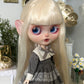 Doll with long blonde hair and blue eyes wearing a gray dress in a decorative setting.